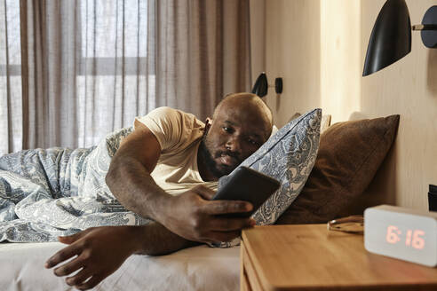 Junger Mann überprüft sein Smartphone auf dem Bett liegend zu Hause - DSHF00136
