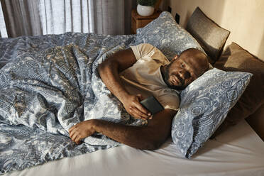 Mann mit Smartphone morgens auf dem Bett liegend zu Hause - DSHF00137