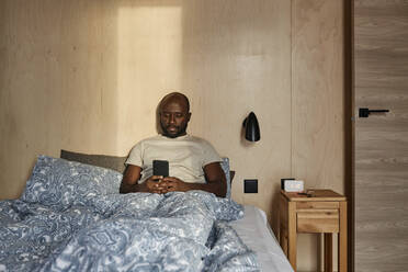 Young man using smart phone sitting on bed in bedroom at home - DSHF00138