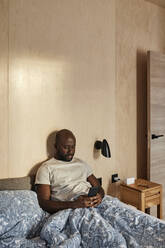 Junger Mann mit Handy auf dem Bett sitzend zu Hause - DSHF00139