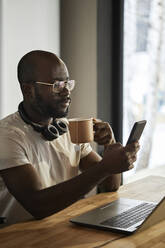 Man using smart phone holding tea cup sitting at home - DSHF00155