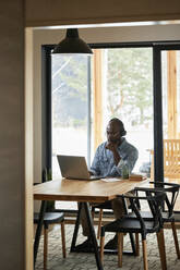 Businessman with hand on chin using laptop on dining table at home - DSHF00167