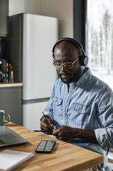 Mann mit Kopfhörern spielt Rhythmus mit Bleistiften auf dem Esstisch zu Hause - DSHF00173