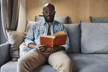 Man reading novel sitting on sofa at home - DSHF00177