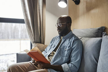Young man reading novel on sofa at home - DSHF00178