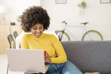 Lächelnde junge Frau, die ein Smartphone benutzt und mit einem Laptop auf dem Sofa im Wohnzimmer sitzt - JCZF00976