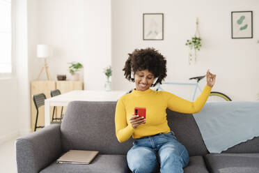 Smiling young woman using smart phone enjoying music through wireless headphones on sofa at home - JCZF00978