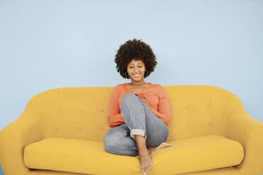 Glückliche junge Frau, die mit ihrem Smartphone auf einem gelben Sofa vor blauem Hintergrund im Internet surft - JCZF00998
