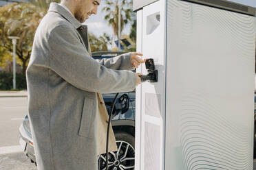 Junger Mann beim Einstecken des Autoladegeräts an der Ladestation - GMCF00258