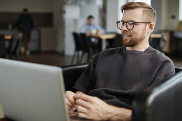 Smiling businessman with eyeglasses sitting with laptop in office - DSHF00208