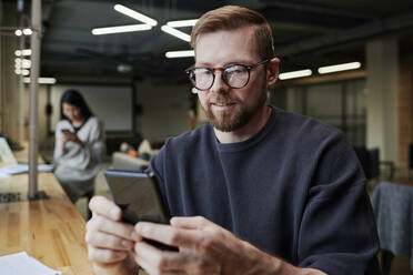 Lächelnder Geschäftsmann mit Brille, der ein Smartphone im Büro benutzt - DSHF00213