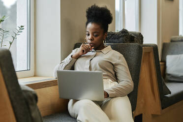 Nachdenkliche Geschäftsfrau mit Hand am Kinn, die auf einen Laptop schaut, der im Büro steht - DSHF00218