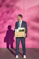 Businessman carrying box with personal belongings in front of pink wall - VEGF05454