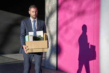 Businessman carrying box of personal belongings by wall on footpath - VEGF05461