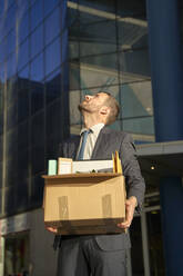 Businessman with box looking up by office building on sunny day - VEGF05463