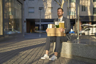 Lächelnder Geschäftsmann mit Kiste auf einer Bank am Fußweg sitzend - VEGF05466