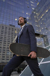 Nachdenklicher Geschäftsmann mit Skateboard geht an einem Bürogebäude vorbei - VEGF05493