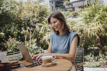 Smiling working woman using laptop in yard - MFF08740