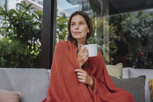 Nachdenkliche Frau mit Kaffeetasse in Decke eingewickelt im Hof - MFF08758