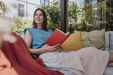Thoughtful woman with book sitting on sofa - MFF08765