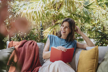 Happy woman with book sitting on sofa at back yard - MFF08771