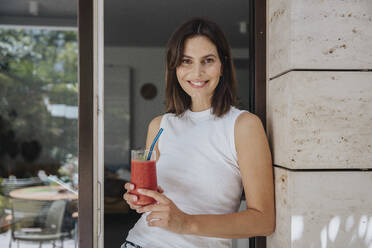 Woman holding smoothie leaning at doorway - MFF08791