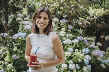 Smiling woman with smoothie glass standing in garden - MFF08793