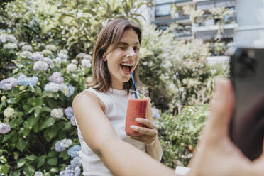 Woman winking holding smoothie glass taking selfie in garden - MFF08796