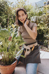 Glückliche Gärtnerin mit Hand im Haar, die neben einer Topfpflanze kniet - MFF08817