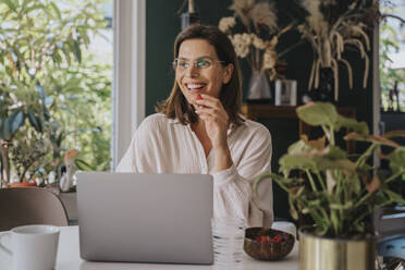 Smiling freelancer with laptop eating strawberry working at home office - MFF08822