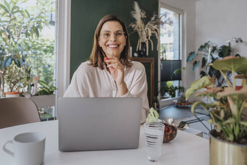 Smiling freelancer with strawberry sitting with laptop at desk in home office - MFF08824