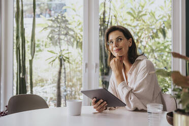 Thoughtful woman with hand on chin holding tablet PC sitting at home - MFF08828