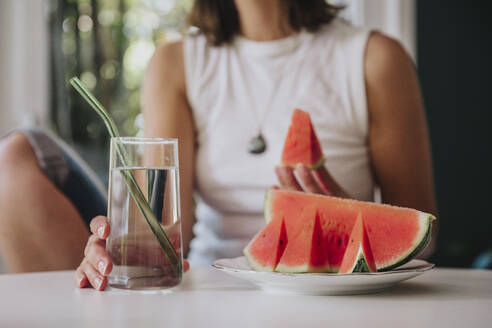 Frau hält Trinkglas an Wassermelonenscheibe in Teller auf Tisch zu Hause - MFF08844