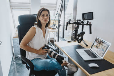 Smiling photographer with digital camera sitting on chair with laptop on table at studio - MFF08864