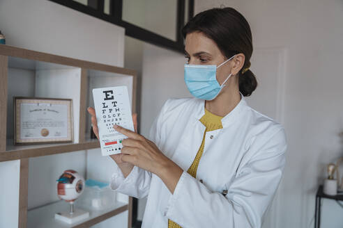 Doctor with protective face mask showing eye chart in clinic - MFF08872