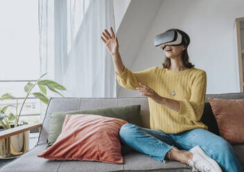Frau mit Virtual-Reality-Headset sitzt zu Hause auf dem Sofa - MFF08894