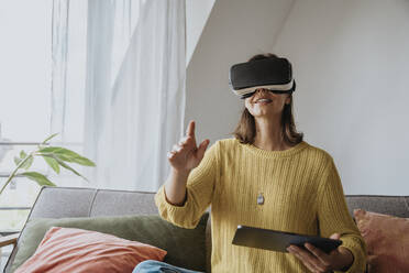 Woman with virtual reality headset holding tablet PC sitting on sofa at home - MFF08896