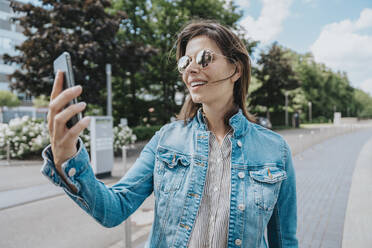 Woman taking selfie through smart phone wearing sunglasses standing at footpath - MFF08911