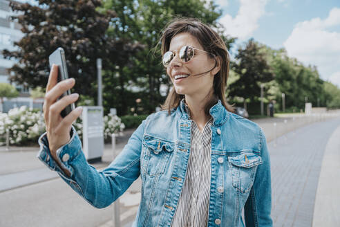 Frau nimmt Selfie durch Smartphone mit Sonnenbrille stehend am Fußweg - MFF08911