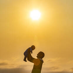 Profile of father holding baby son (6-11 months) aloft against yellow sky at sunset - TETF01373