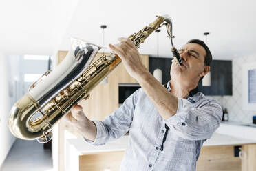 Musician with eyes closed blowing saxophone practicing in kitchen at home - JRFF05341