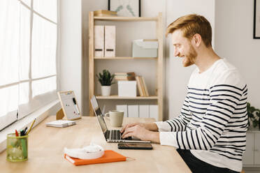 Smiling young man typing on laptop sitting at table in living room - XLGF02861