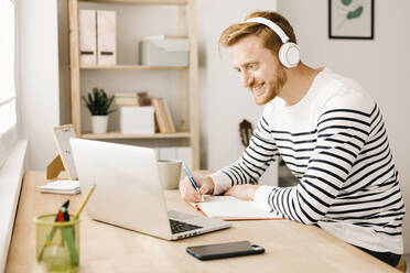 Smiling man wearing wireless headphones looking at laptop in living room - XLGF02864