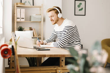 Man listening music through wireless headphones sitting with laptop in living room - XLGF02866