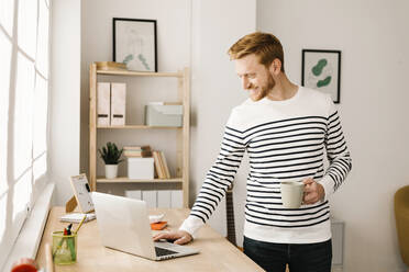 Glücklicher junger Mann mit Kaffeetasse und Laptop am Wohnzimmertisch stehend - XLGF02867