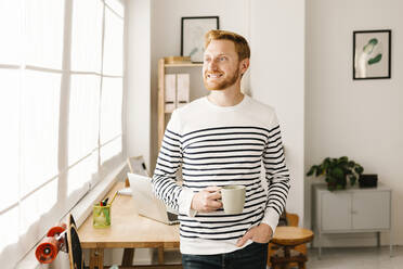 Lächelnder junger Mann mit Kaffeetasse und Hand in der Tasche zu Hause stehend - XLGF02868