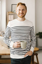 Lächelnder Mann mit Hand in der Tasche, der eine Kaffeetasse hält, steht zu Hause - XLGF02869