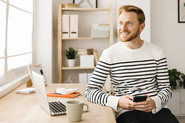 Glücklicher junger Mann mit Smartphone, Laptop und Kaffeetasse am Tisch - XLGF02872