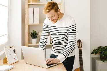 Lächelnder Mann mit braunen Haaren, der einen Laptop benutzt und am Tisch im Wohnzimmer steht - XLGF02873