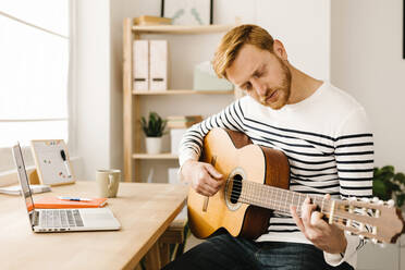 Young man practicing guitar sitting at table in living room - XLGF02874